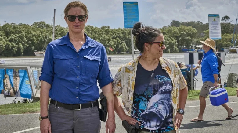 Kate Box as Dulcie in blue shirt and grey pants next to Madeleine Sami as Eddie in fish t-shirt and Hawaiian shirt in front of marina in Deadloch