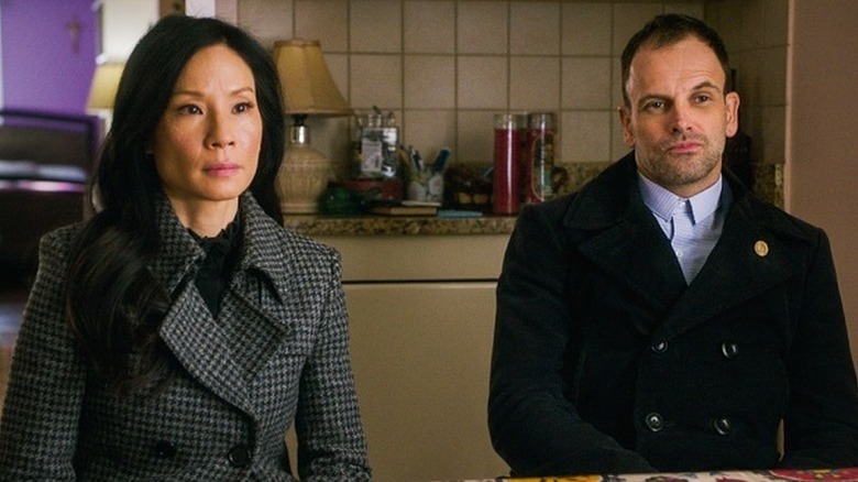 Lucy Liu as Joan Watson sitting in checkered grey coat net to Jonny Lee Miller as Sherlock Holmes in black coat in Elementary