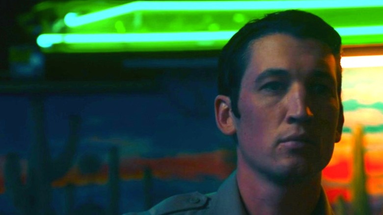 Miles Teller as Martin in sheriff's uniform standing in front of green neon and desert backdrop in Too Old to Die Young