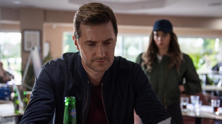 Adam Price (Richard Armitage) reads a letter while a woman (Hannah John-Kamen) approaches him from behind in The Stranger