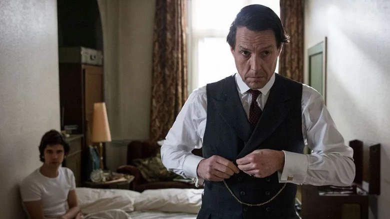 Hugh Grant as Jeremy Thorpe buttoning his suit in front of bed where Ben Whishaw as Norman Scott sits in A Very English Scandal