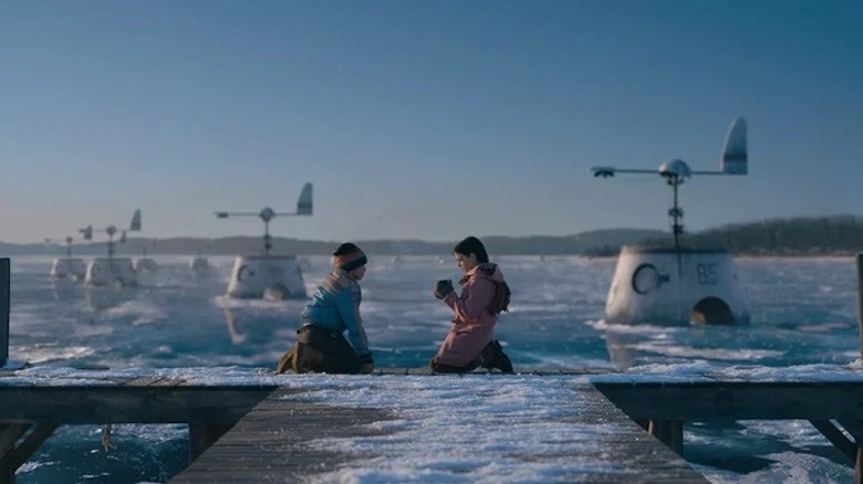 Two children kneeling on dock by icy lake with metal structures behind them in Tales From the Loop