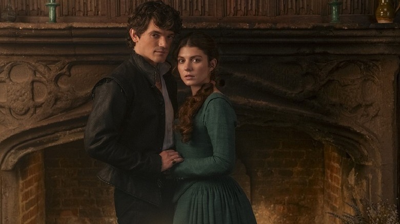 Edward Bluemel as Guildfordholding hands with Emily Bader as Jane in period clothing in front of large fireplace in My Lady Jane