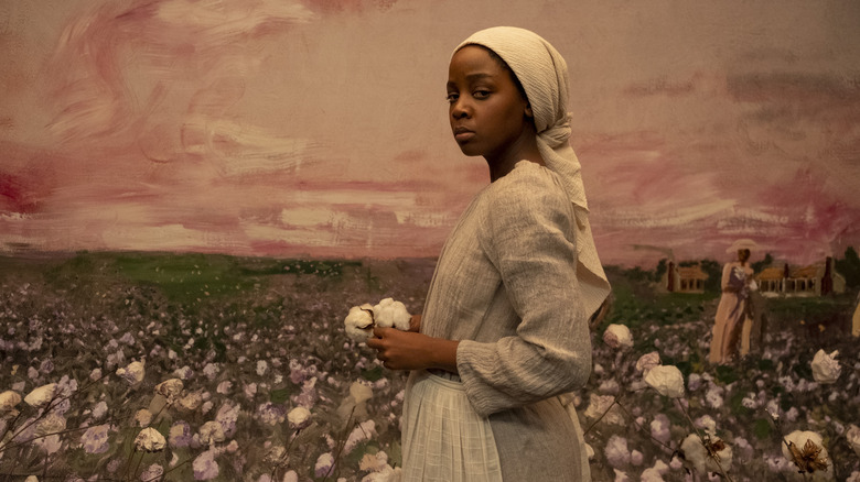 Thuso Mbedu as Cora in grey/white dress and head scarf in front of painted backdrop of cotton fields in The Underground Railroad