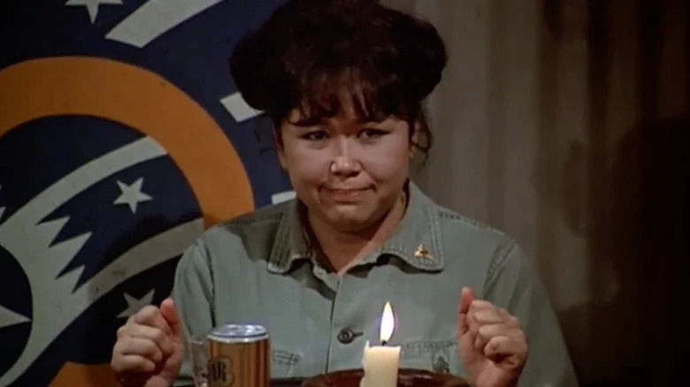 Kellye Yamato (Kellye Nakahara) sits at a table with a beer in M*A*S*H