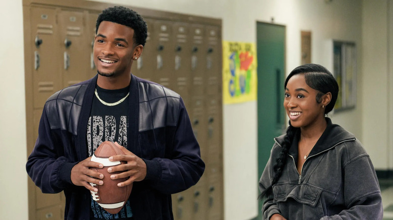 KJ Jeremy (Nathaniel McIntyre) holds a football in a high school hallway smiling with Amina (Alexis Chikaeze) in All American