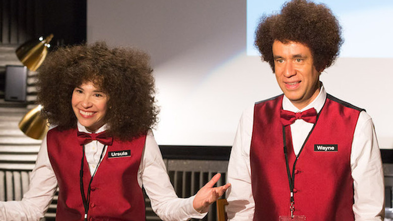 Ursula (Carrie Brownstein) and Wayne (Fred Armisen) stand in front of a projection screen in Portlandia