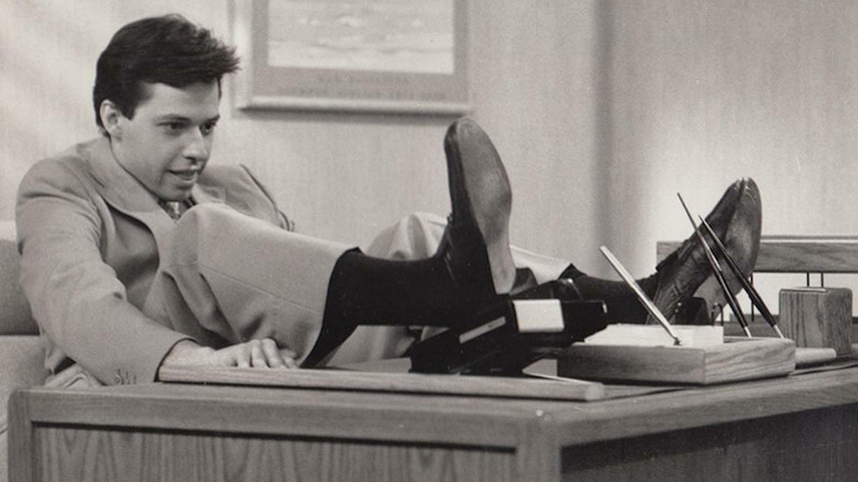 John Cryer in black and white as Teddy sitting awkwardly with feet up on desk in The Famous Teddy Z