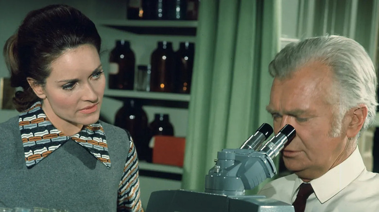 Betty Jones (Lee Meriwether) speaks to Barnaby Jones (Buddy Ebsen) as he looks into a microscope in Barnaby Jones