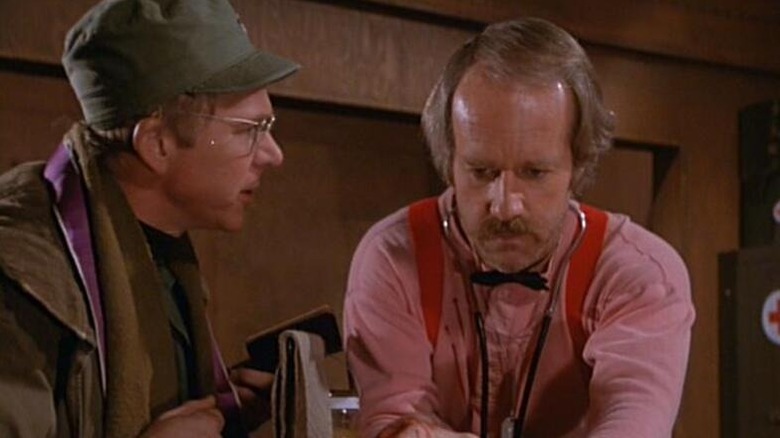 John Mulcahy (William Christopher) speaks to B.J. Hunnicut (Mike Farrell) as he works intensely in M*A*S*H