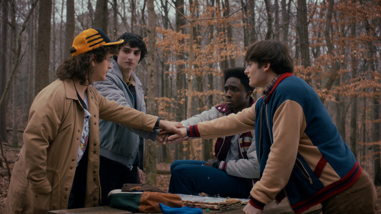 Dustin Henderson (Gaten Matarazzo), Mike Wheeler (Finn Wolfhard), Lucas Sinclair (Caleb McLaughlin), and Will Byers (Noah Schnapp) put their hands together in a forest in Stranger Things