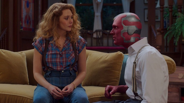 Wanda crying on a couch while Vision wearing normal clothes kneels next to her