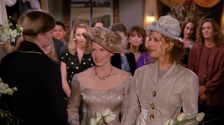 Jane Sibbett as Carol in tan dress and hat standing next to Jessica Hecht as Susan in suit and hat at wedding ceremony with Lisa Kudrow as Phoebe and others behind them in Friends