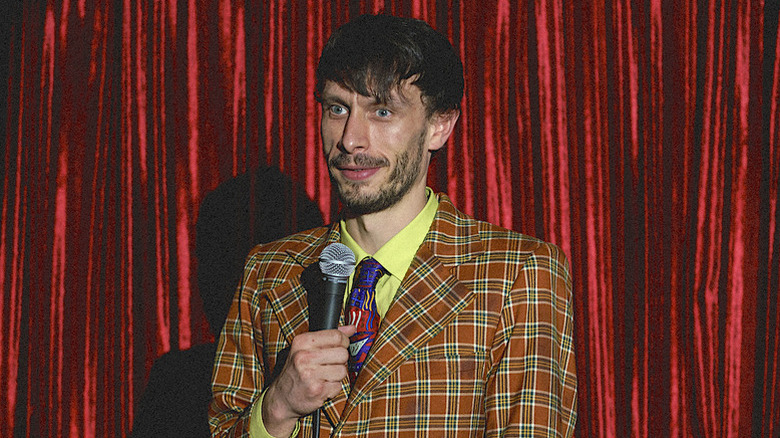 Richard Gadd as Donny in plaid suit in front of red curtain holding microphone in Baby Reindeer