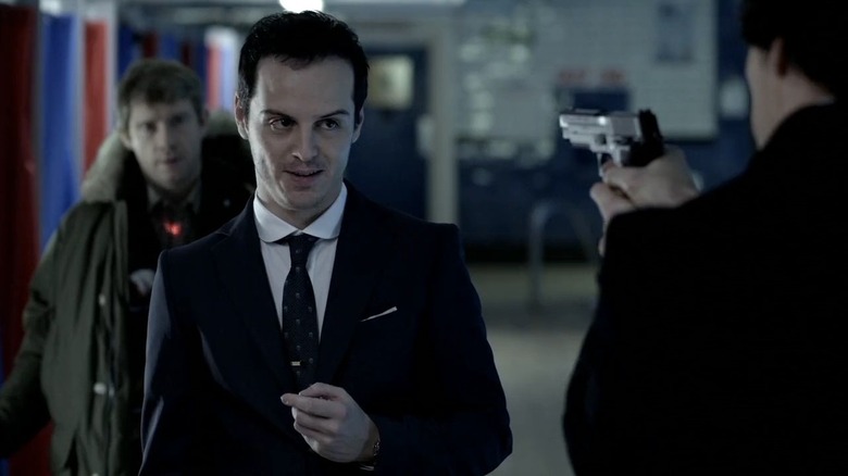 Andrew Scott as Moriarty smiling at Benedict Cumberbatch as Sherlock holding a gun while Martin Freeman as Watson stands behind him