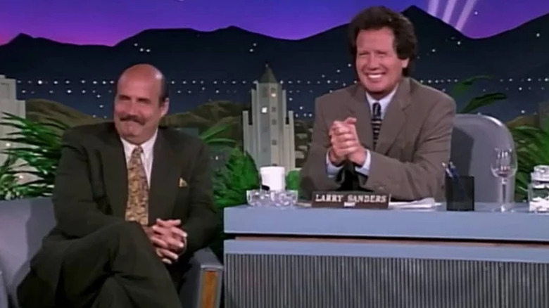 Larry Sanders (Garry Shandling) sits happily at his talk show desk, with Hank Kingsley (Jeffrey Tambor) seated next to him in The Larry Sanders Show