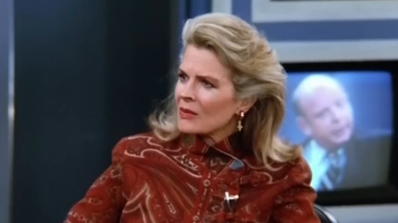 Murphy Brown sitting with a stern expression while a screen behind her shows Stuart Best on Murphy Brown S7E13