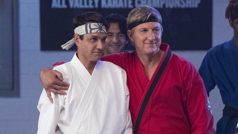 Johnny Lawrence (William Zabka) puts his arm around Daniel LaRusso (Ralph Macchio) as they both wear karate gis on Cobra Kai