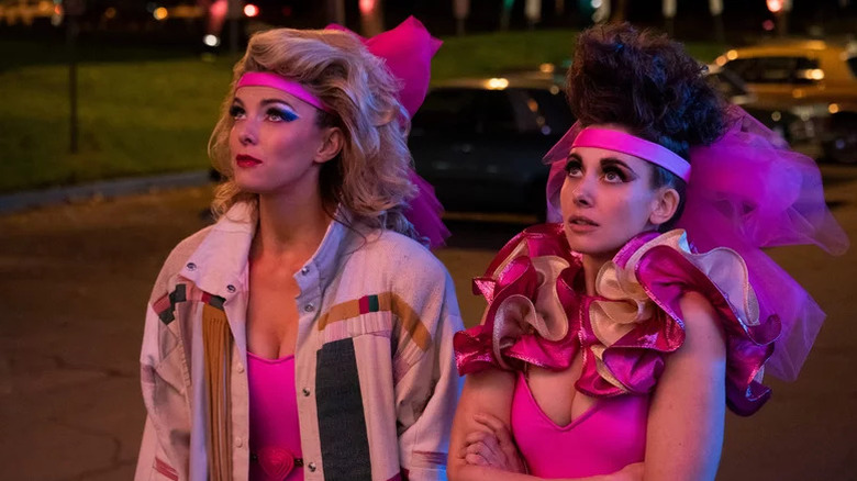 Debbie Eagan (Betty Gilpin) and Ruth Wilder (Alison Brie) wear flamboyant wrestling outfits and makeup while looking up in a parking lot on GLOW