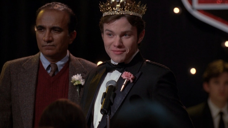 Kurt wearing tux and crown in front of microphone next to Principal Figgins