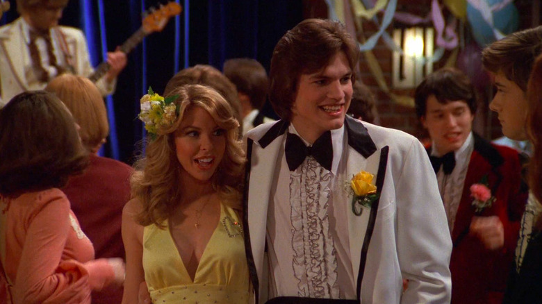 Pam in yellow dress standing next to Kelso in white tux, both smiling