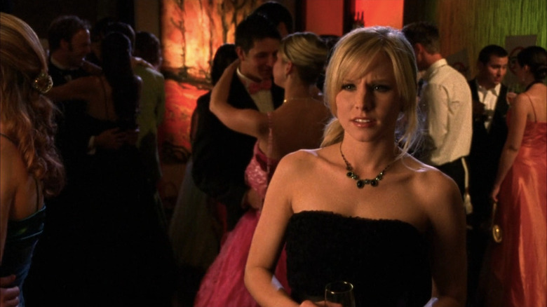 Veronica in a black dress surrounded by prom attendees, looking confused
