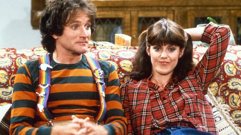 Robin Williams as Mork in striped shirt and suspenders sitting next to Pam Dawber as Mindy on the couch in Mork & Mindy