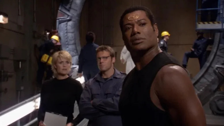 "Christopher Judge as Teal'c with golden circle on his forehand standing in front of Amanda Tapping as Samantha Carter and Michael Shanks and Daniel Jackson with large metal circle behind them on Stargate SG-1