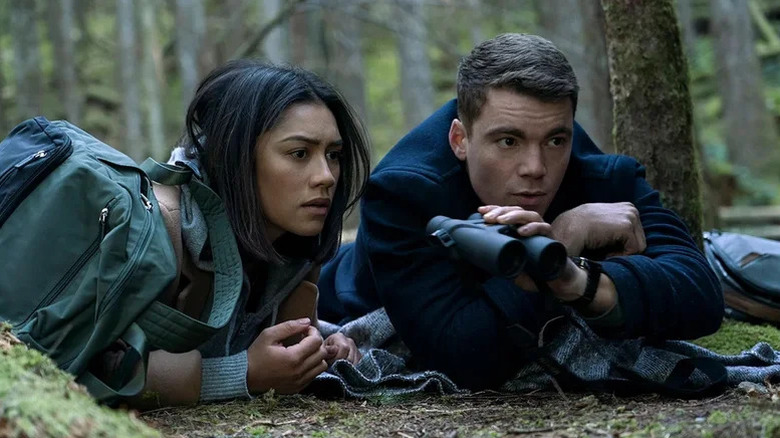 Luciane Buchanan as Rose laying on stomach on forest floor next to Gabriel Basso as Peter holding binoculars in The Night Agent