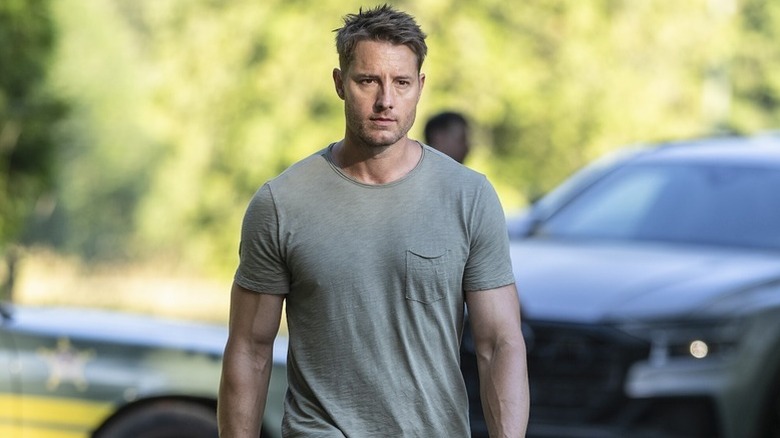 Justin Hartley as Colter in t-shirt walking in front of cars in the background in Tracker