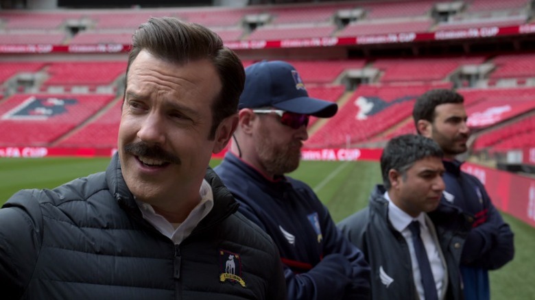 Ted smiling and looking to his right while standing next to Coach Beard, Nate, and Roy on Ted Lasso S2E8