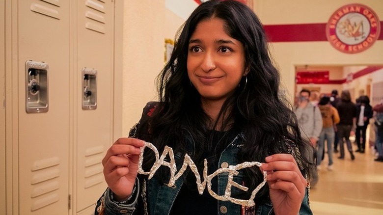 Maitreyi Ramakrishnan as Devi standing in school hallway holding up the word 'DANCE' in Never Have I Ever