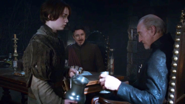 Maisie Williams as Arya serving wine to Charles Dance as Tywin Lannister while Aiden Gillan as Littlefinger sits and watches in Game of Thrones
