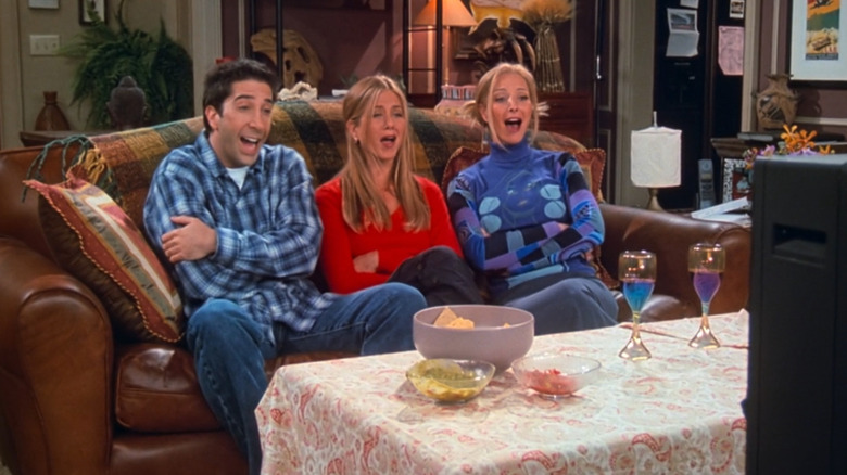 David Schwimmer as Ross Geller, Jennifer Aniston as Rachel Green, and Lisa Kudrow as Phoebe Buffay sitting on Ross' couch and laughing at the tv while the Pottery Barn Apothecary table is hidden under a sheet in Friends
