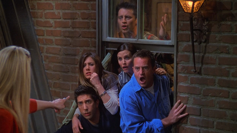Lisa Kudrow as Phoebe Buffay holding the lottery tickets hostage on the balcony while David Schwimmer as Ross Geller, Jennifer Aniston as Rachel Green, Courteney Cox as Monica Geller, Matt LeBlanc as Joey Tribbiani, and Matthew Perry as Chandler Bing watch through the window in horror in Friends