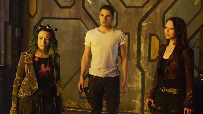 Jodelle Ferland's Emily Kolburn, Mark Bendavid's Derrick Moss, and Melissa O'Neil's Portia Lin beside a large metal wall on Dark Matter