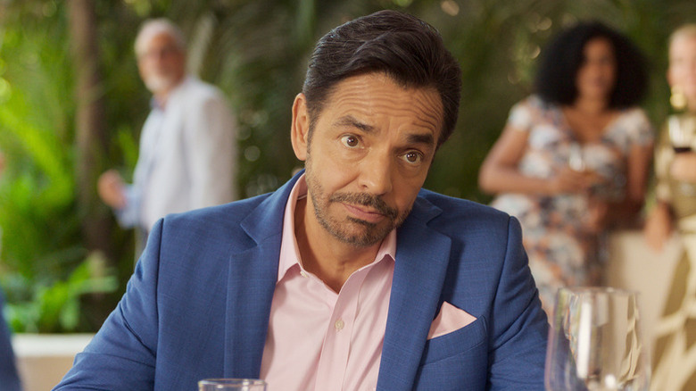 Eugenio Derbez as Maximo sitting at restaurant in blue and pink suit in Acapulco