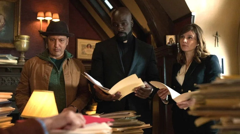 Ben, David in priest uniform, and Kristen standing in room filled with stacks of papers in Evil