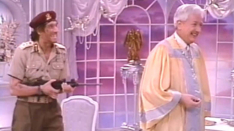 Actor portraying Muammar Gaddafi pointing a machine gun at Joseph Maher as Saint Peter in Second Chances