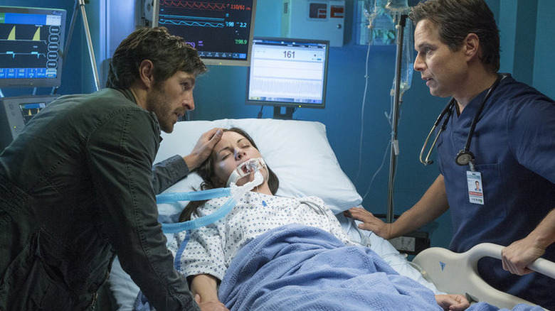 Eoin Macken as T.C. with hand on the forehead of Jill Flint as Jordan, in hospital bed on ventilator, next to Scott Wolf as Scott in "Night Shift."