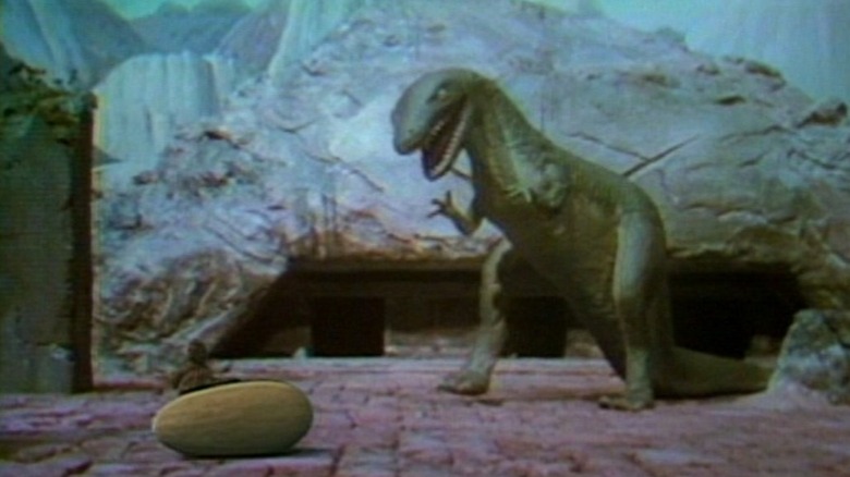 Grumpy the Tyrannosaurus standing over a large oval egg on a rocky terrain