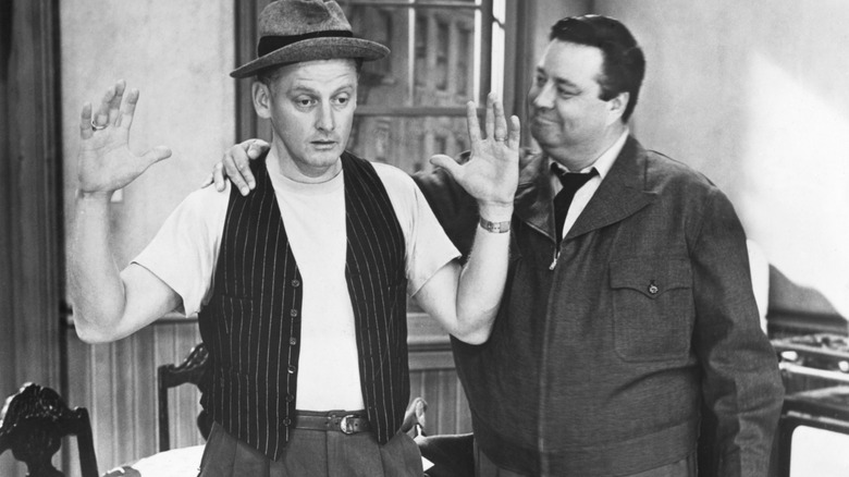 Ed Norton with hands in the air while Ralph Kramden smiles and puts arm around him