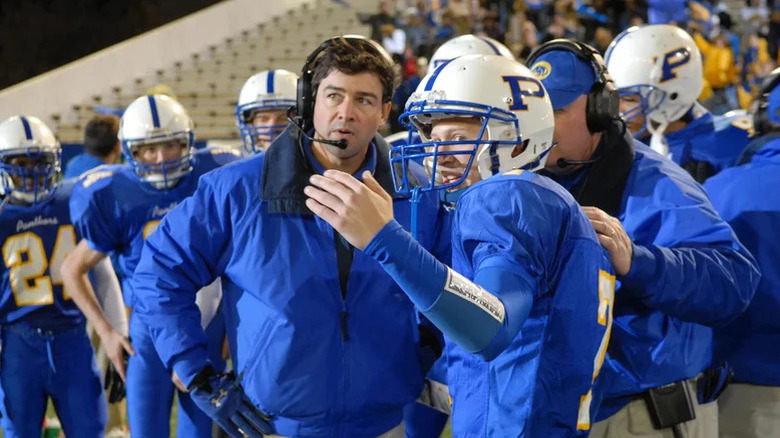 Kyle Chandler as Coach Taylor in blue jacket with headset talking to Zach Gilford as Matt in blue football uniform in Friday Night Lights