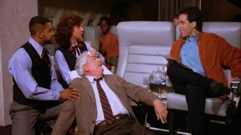 Jerry Seinfeld as himself sitting on a plane looking at Joseph Maher as Gavin being taken care of by flight attendants in Seinfeld