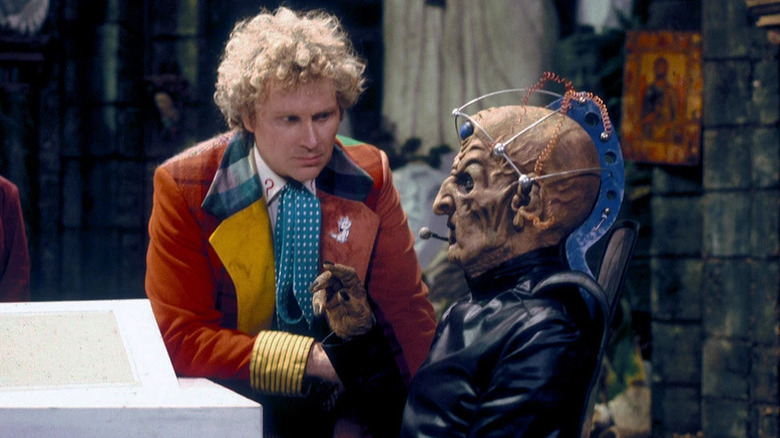 The Sixth Doctor (Colin Baker) speaks to a seated alien in Doctor Who