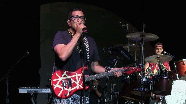 Sal holding a guitar and speaking into a microphone while Joe sits at the drumset on Impractical Jokers S2E23