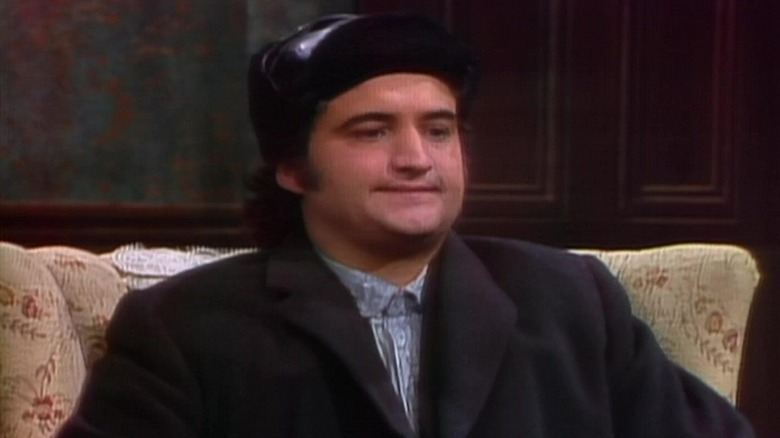 John Belushi as a foreign man sitting in a chair in the first episode of SNL