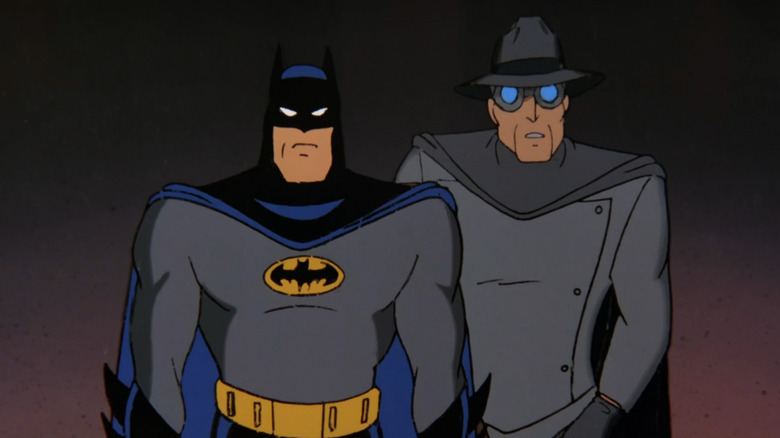Batman and the Gray Ghost standing side by side. On the left, Batman is wearing a gray costume with a black and blue cape and cowl with two pointed ears. He has a yellow and black bat symbol on his chest, and a yellow utility belt around his waist. On the right, is an older Gray Ghost wearing a light gray costume with dark gray cape, a gray trilby, and gray goggles. They're both looking at the camera.