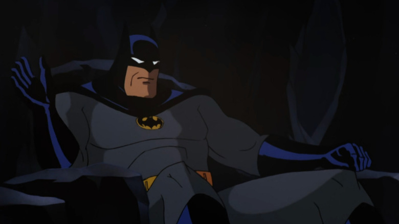 Batman is sitting down in a rocky chair. He's wearing a gray costume with a black and blue cape and cowl with two pointed ears. He has a yellow and black bat symbol on his chest. He also has black and blue gloves.