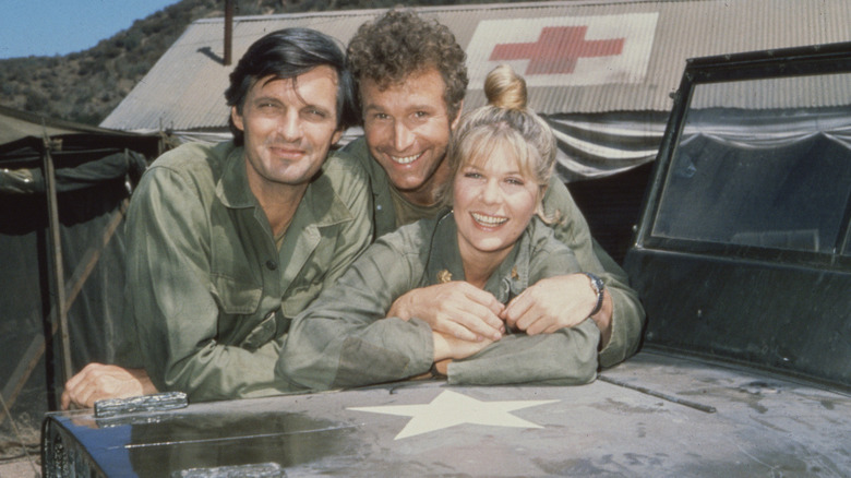 Alan Alda, Wayne Rogers, and Loretta Swift all smiling and leaning on a van